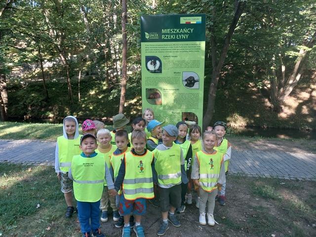 Dzieci na tle zielonych drzew oraz tablicy informującej o mieszkańcach rzeki Łyny