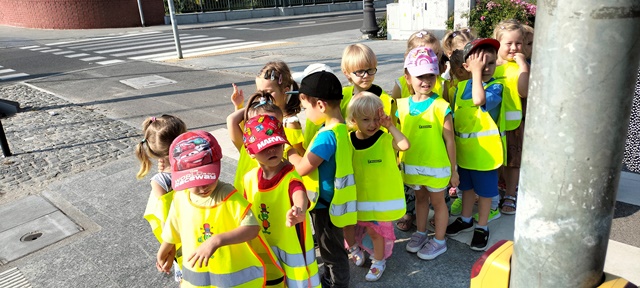 Grupa dzieci w odblaskowych kamizelkach stoi parami przed przejściem dla pieszych.