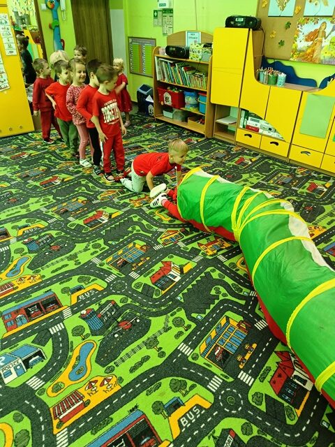 Na dywanie rozłożony jest tunel, w który wchodzą dzieci.