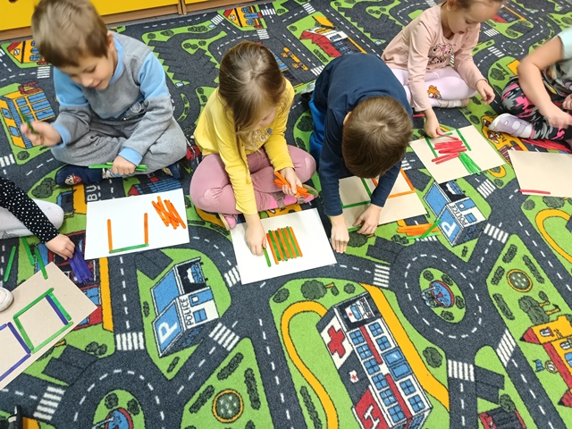 Dzieci na dywanie na rozłożonych kartkach przeliczają patyczki.