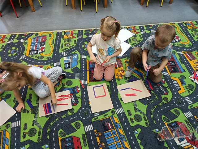 Dzieci na dywanie układają na kartkach dowolne kompozycje z drewnianych kolorowych patyczków.