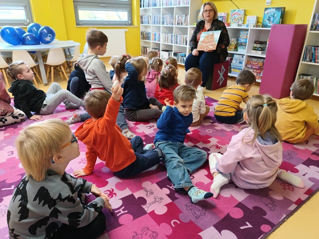 Dzieci siedzą na dywanie i słuchają opowiadania czytanego przez panią z biblioteki oraz oglądają ilustrację. 