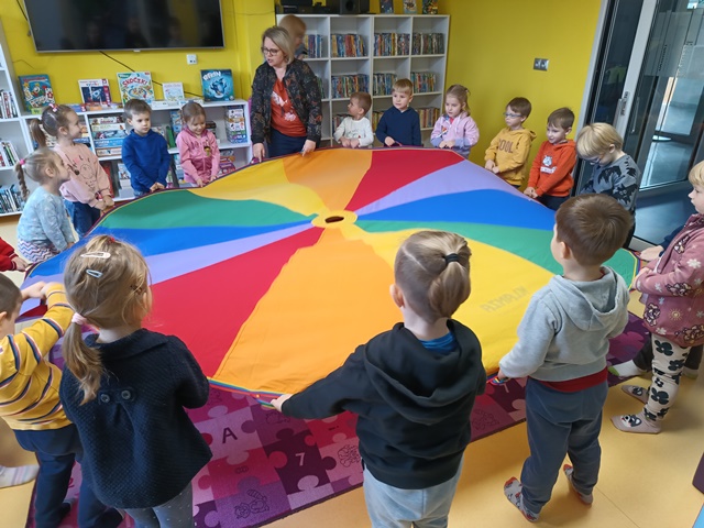 Grupa dzieci wraz z panią bibliotekarką trzyma kolorowa chustę animacyjną. 