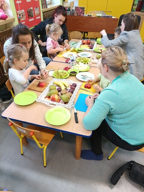Przy stole siedzą mamy i dzieci. Mamy kroją na kawałki owoce, a dzieci wbijają wykałaczki w połówki jabłek. 