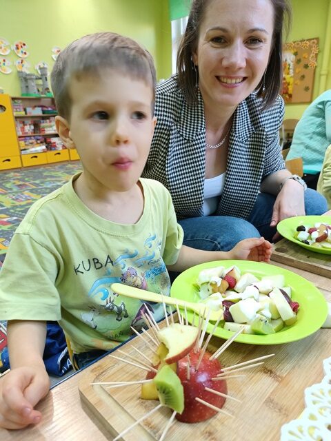 Mama z synkiem siedzą przy stoliku. Chłopiec zjada łyżeczką owoce.