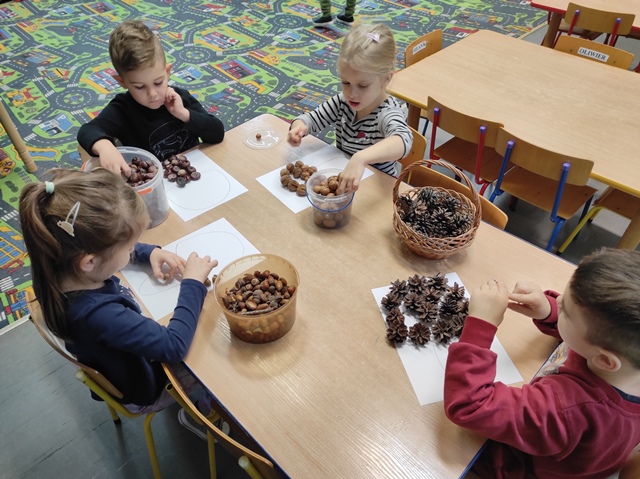 Dzieci przy stoliku układają dary natury na kartce tworząc różne kształty 