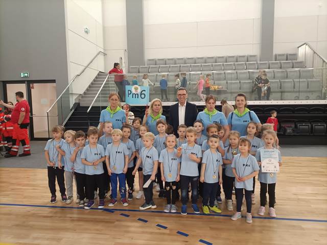 Grupa dzieci w błękitnych koszulkach, stoi uhonorowana medalami wraz z 4 paniami i prezydentem Olsztyna Piotrem Grzymowiczem.
