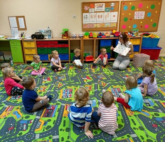 Dzieci siedzą na dywanie w kręgu w siadzie skrzyżnym, a nauczyciel czyta książkę o kolorowych kropkach. 