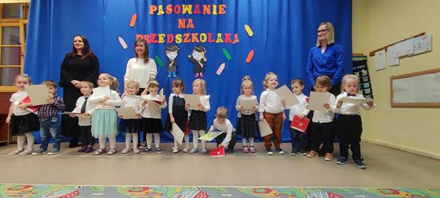 Dzieci oraz Panie stoją na tle dekoracji z okazji Pasowania. Przedszkolaki trzymają w ręku dyplomy oraz upominki – książeczki. 