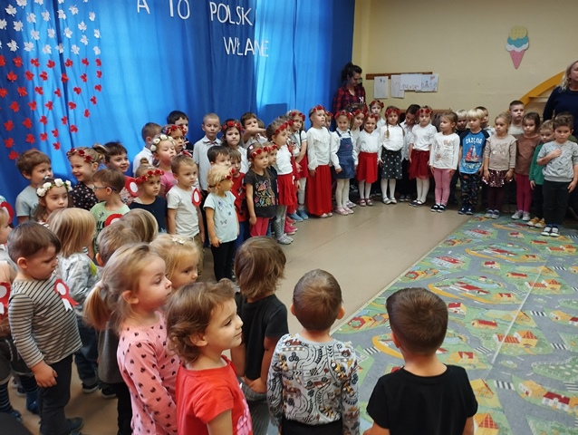 Dzieci stoją w półkolu na tle dekoracji z okazji 11 listopada i śpiewają hymn. 