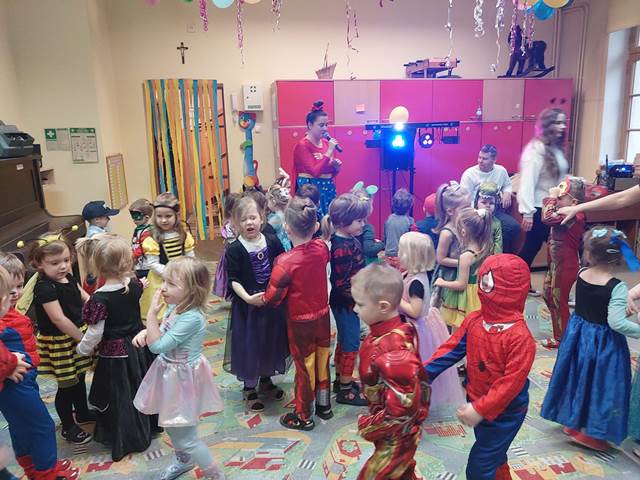 Przebrane dzieciaki tańczą przy muzyce. Sala jest ozdobiona balonami i serpentynami. 