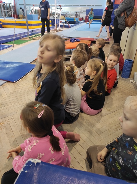 Dzieci siedzą w sali gimnastycznej, przyglądają się ćwiczeniom gimnastyczek