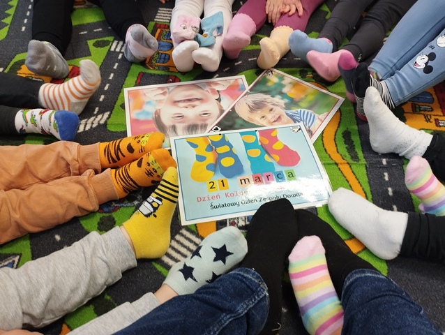 Na dywanie leżą ilustracje przedstawiające Dzień Kolorowej Skarpetki symbolizujący Dzień osób z Zespołem Downa. Wokół znajdują się stopy dzieci ubrane w kolorowe skarpetki.