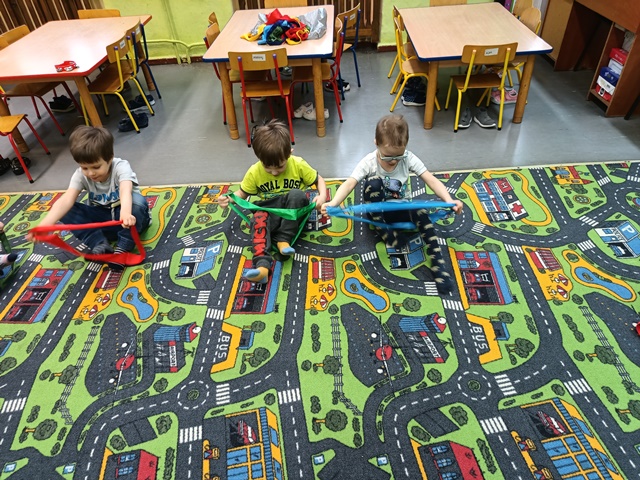  Trzech chłopców siedzi na dywanie trzymając przed sobą oburącz szarfę. Próbują włożyć dwie nogi do szarfy.