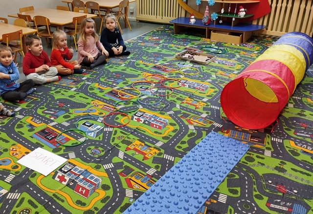 Przed siedzącymi na dywanie dziećmi  rozłożona jest ścieżka sensoryczna, która będą pokonywać w skarpetkach. 
