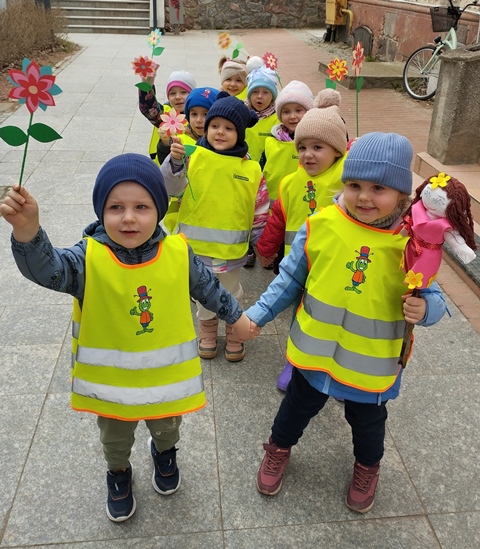 Dzieci ubrane w kamizelki odblaskowe stoją w parach. Dziecko w pierwszej parze trzyma w ręku Marzannę, pozostałe kolorowe kwiatki wykonane z bibuły. 