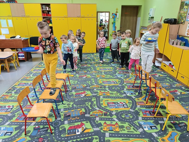 Dzieci ustawione są w dwóch rzędach, przed którymi stoją krzesła. Dzieci przechodzą po krzesłach, trzymając w dłoni ringo z małą piłką. 