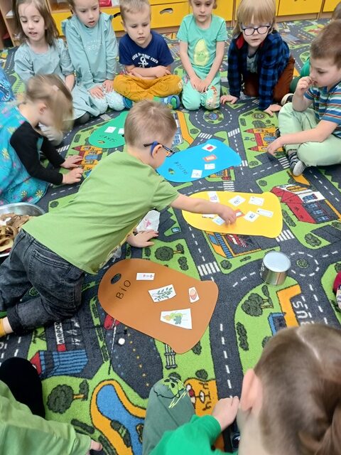 Dzieci siedzą na dywanie, na którym rozłożone są sylwety koszy na śmieci w kolorach: brązowy, żółty, niebieski, zielony. Dzieci dopasowują obrazek przedstawiający konkretny śmieć do koloru kosza.