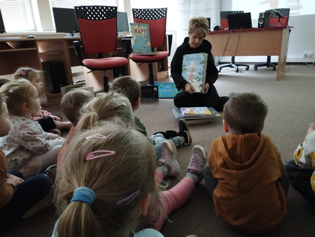 Przedszkolaki siedzą w bibliotece na dywanie. Pani prezentuje dzieciom różne książki.