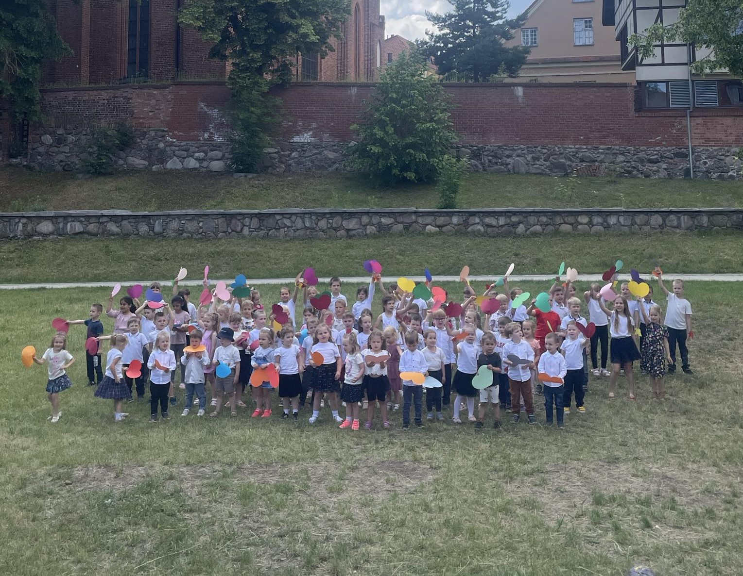 Dzieci ze wszystkich grup stoją na trawie i śpiewają piosenkę dla rodziców. W ręku trzymają kolorowe papierowe serca.