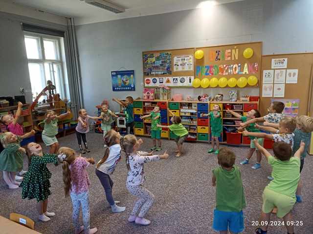 Dzieci ustawione w kole tańczą do piosenki "Ciu ciu ła". W tle na tablicy wisi napis i balony