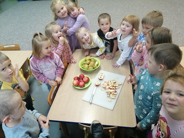 Dzieci stoją wokół stolika na którym znajdują się jabłka i gruszki przeznaczone do degustacji.  