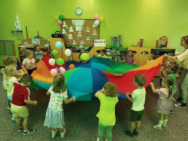 Przedszkolaki trzymają w kolorową chustę. Na chuście znajdują się kolorowe balony. Dzieci wachlują chustą tak, aby balony nie wypadł z chusty. 