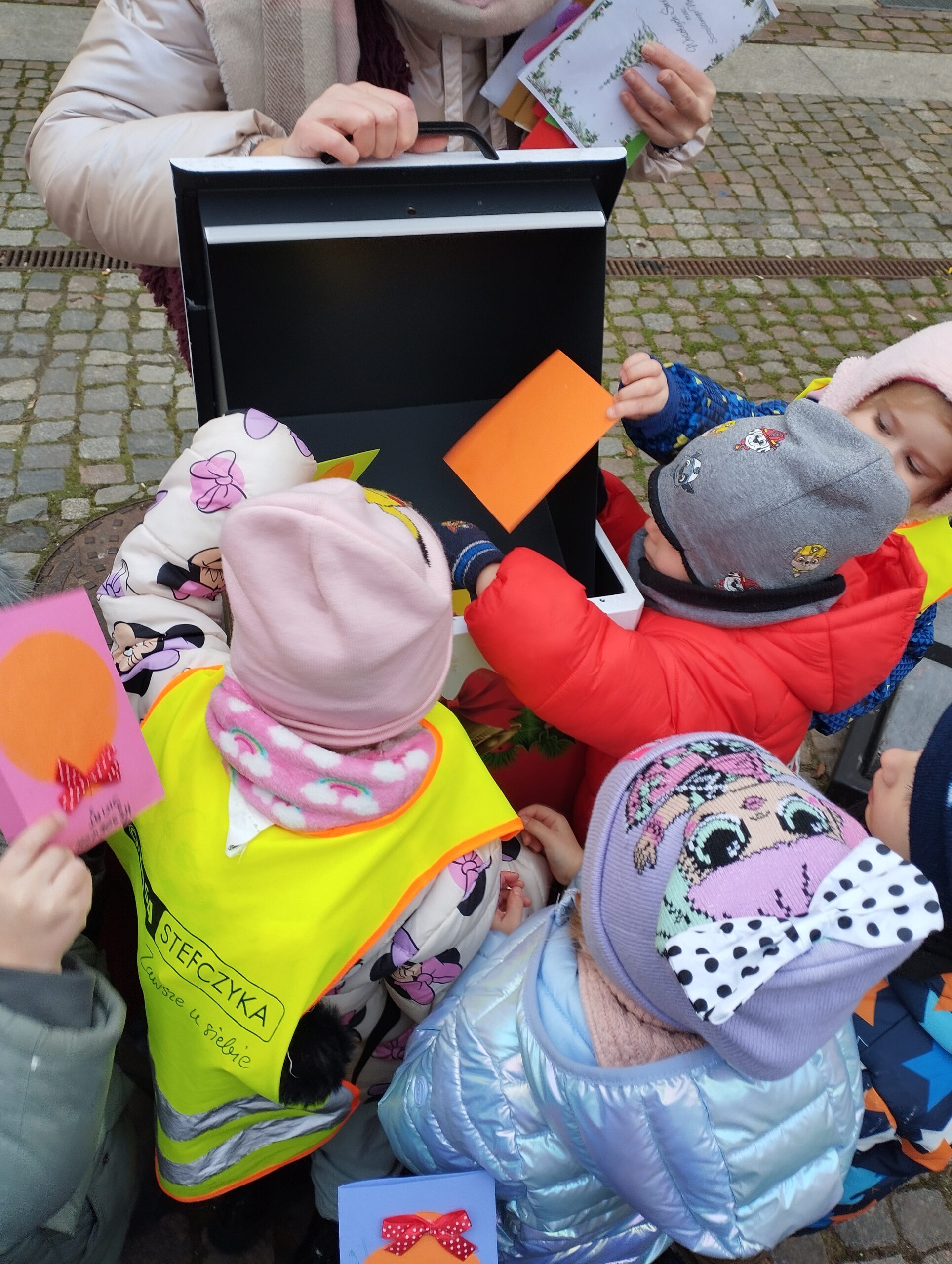 1. Dzieci ubrane w kurtki wrzucają swoje kolorowe kartki do skrzynki umieszczonej przed Biblioteką Wojewódzką. Obok stoi Pani, która otwiera skrzynkę.