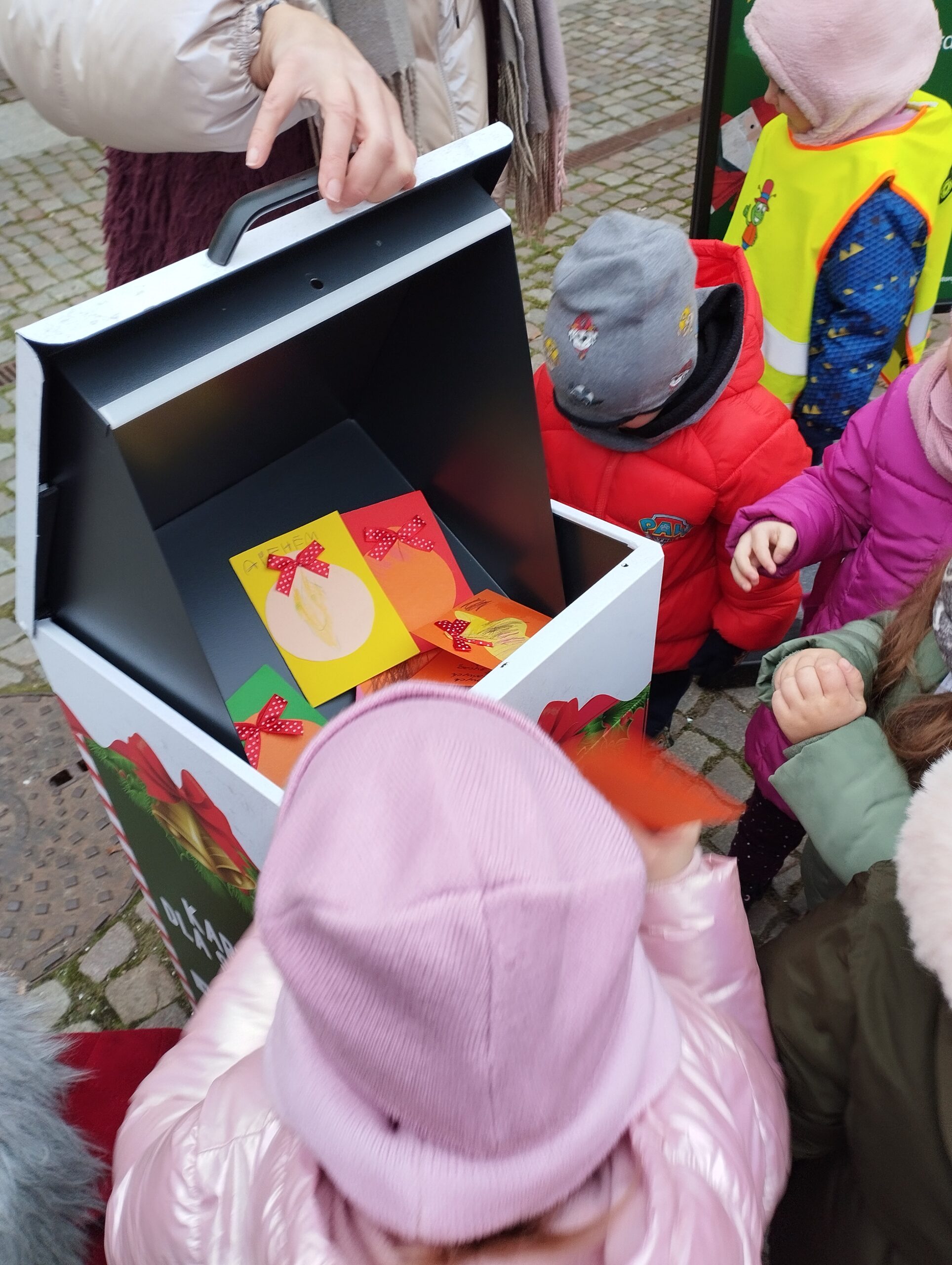 2. Dzieci ubrane w kurtki wrzucają swoje kolorowe kartki do skrzynki umieszczonej przed Biblioteką Wojewódzką. Obok stoi Pani, która otwiera skrzynkę.