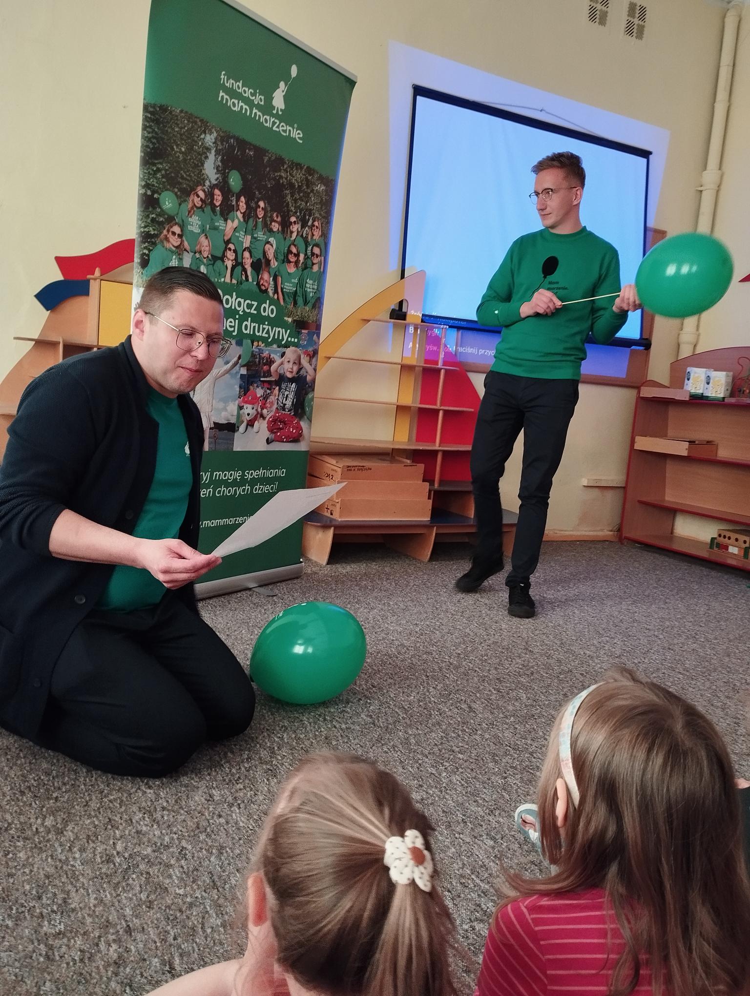  Jeden z wolontariuszy czyta dzieciom wiersz. Wolontariusze trzymają w rękach balony.