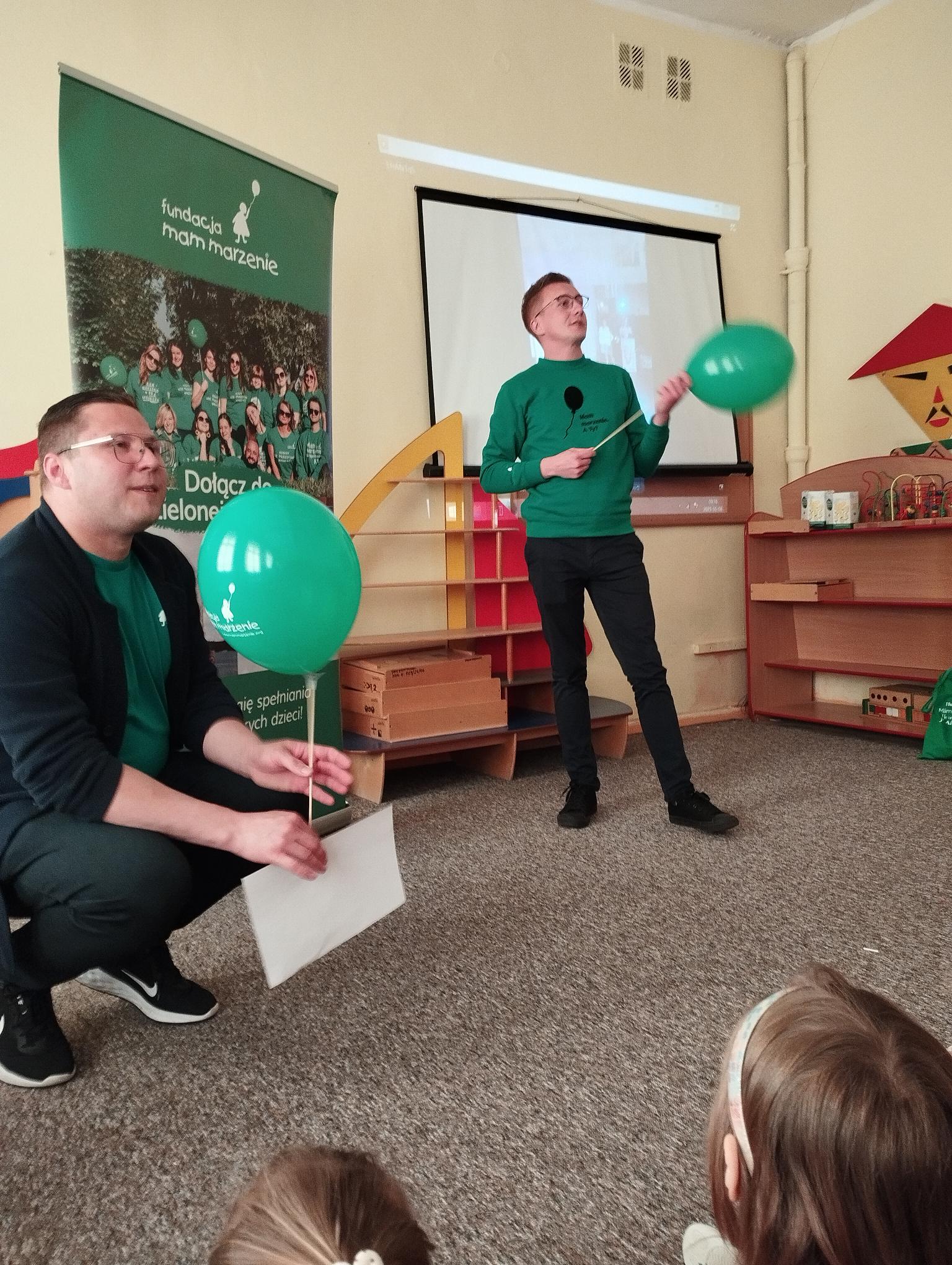 2. Wolontariusze ubrani w zielone koszulki, trzymając balony opowiadają o swoich działaniach.