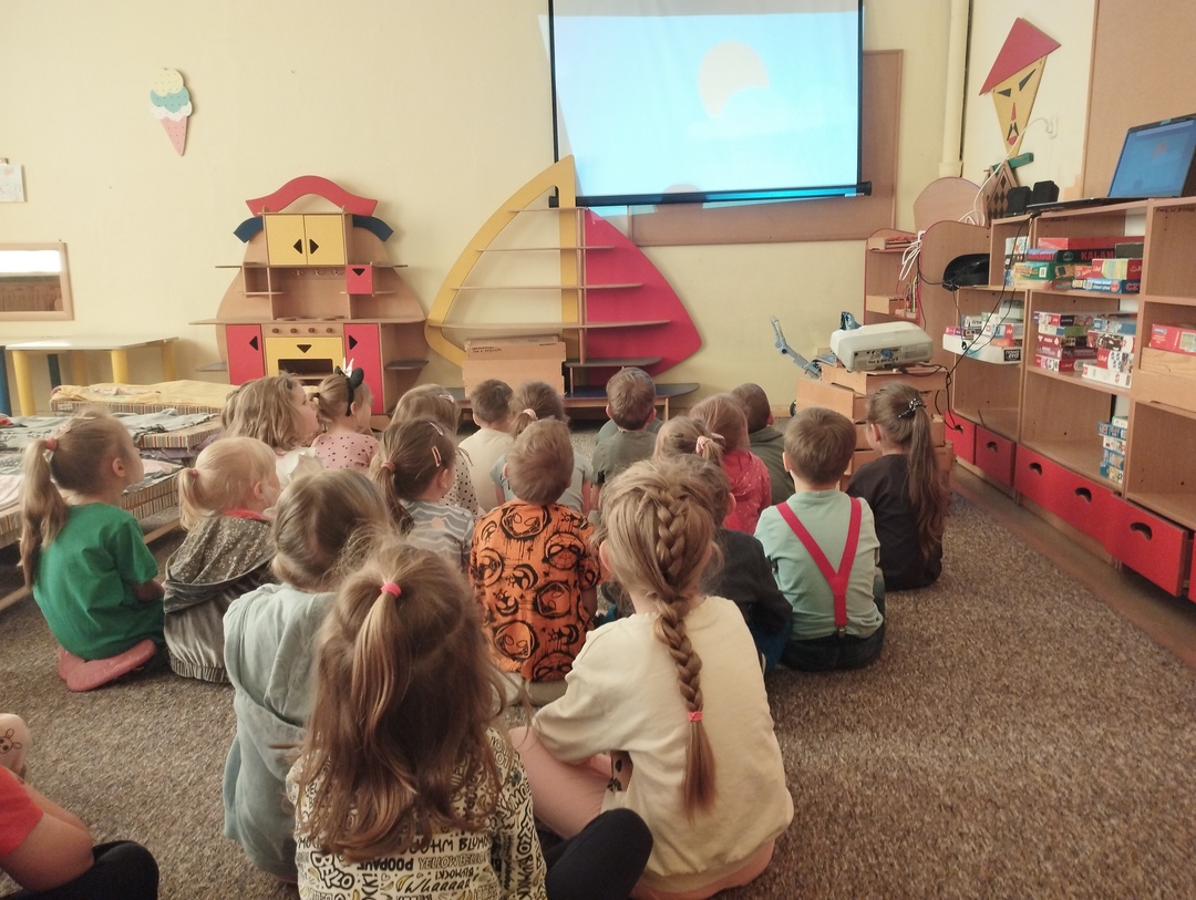 Dzieci siedzą na dywanie w rozsypce ze skierowaną twarzą ku projektorowi. Oglądają film o lecie.